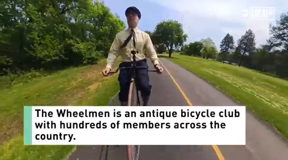 Bill Soloway rides a big-wheel bicycle down a tree-lined paved road. He has light skin, glasses, an old-fashioned cap, a button-up shirt and tie, and capri-length navy old-fashioned pants. A caption reads The Wheelmen is an antique bicycle club with hundreds of members across the country.