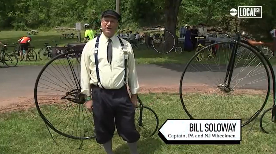 Bill Soloway is interviewed by 6ABC as the caption of the NJ and PA chapters of the Wheelmen with two big-wheel antique bicycles behind him in a public park. Bill wears old-fashioned dress with a buttoned shirt, a tie, suspenders, and black pants to the knee. He has light skin, gray hair, and glasses.