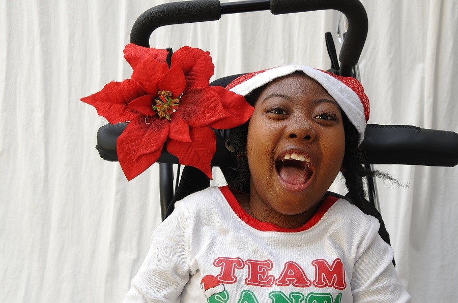 Zoë is radiant as she sits in her black mobility device adorned with a blooming poinsettia. Zoë has brown skin, brown eyes, textured black hair, a Santa holiday hat, and a shirt that reads Team Santa.