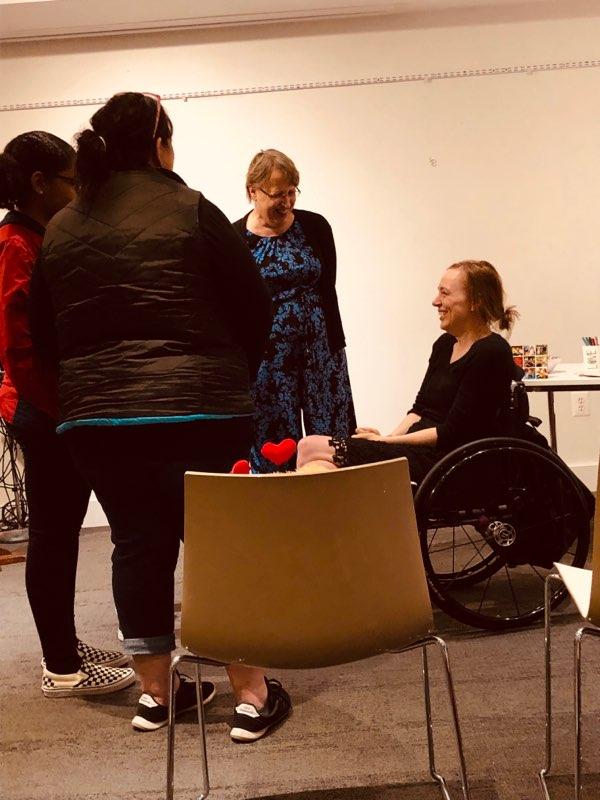 Cindy shares a smile with her daughter, Beth, as they speak to two women in a classroom setting. Cindy has short hair and light skin and wears a black and blue printed pantsuit. Beth has light skin and brown hair pulled back, and she wears all black as she sits in a black manual wheelchair.