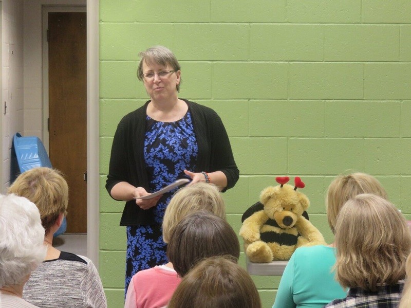 Cindy addresses a group of women in seats. She has light skin, short gray hair, and glasses.