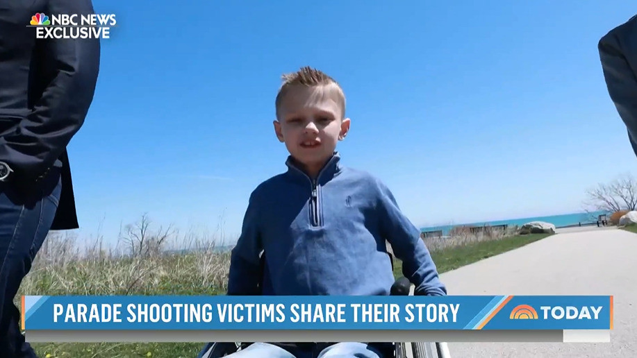 Cooper Roberts is an 8-year-old boy pictured outdoors rolling his manual wheelchair alongside two individuals walking beside him. Cooper has light skin, short blonde hair, and a blue sweater. The news coverage ticker reads NBC News Exclusive: Parade Shooting Victims Share Their Story.