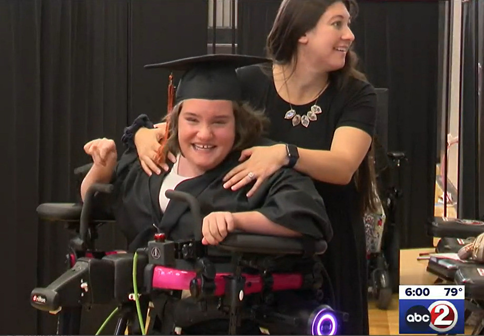 18-year-old Ella Weiske beams in her graduation black cap and gown as she stands in her new Trexo robotic walking device. Ella has light skin, light eyes, and shoulder-length brown hair. Standing behind her, a friend with light skin and long wavy brown hair touches her shoulders.