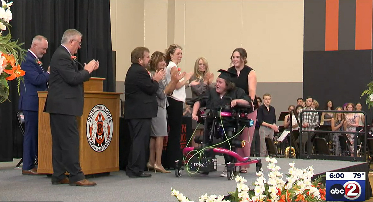 18-year-old Ella Weiske is visibly emotional with joy as she walks across the graduation stage in her Trexo walking device. She has light skin, brown hair, and a black cap and gown with teachers and administrators standing and applauding for her at the podium. She is assisted by a friend in a black dress with light skin and brown hair.