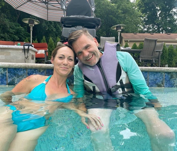 Frank Guirlinger sits in a pool wearing a long-sleeved swim shirt and a gray flotation vest. He has light skin and gray hair. The woman in the pool to his right has brown hair, light skin, and a turquoise bikini.