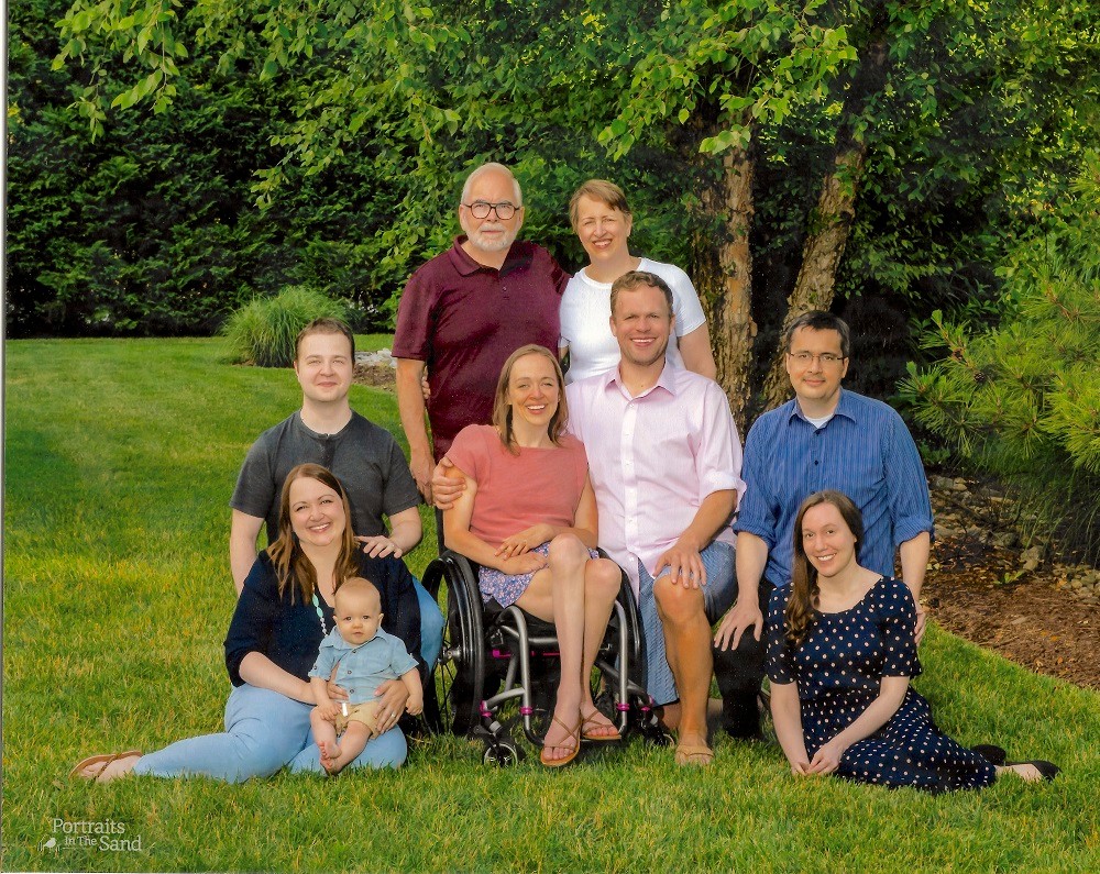 A group portrait of Cindy Kolbe and her family on a lawn. They are dressed in semi-formal spring attire. Cindy is in the back row with light skin, a white blouse, red lipstick, and short sandy brown hair. Her daughter, Beth, is seated in her black manual wheelchair.