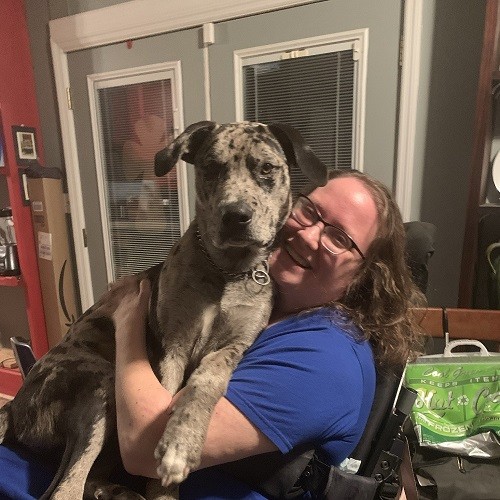 Jennifer Jenkins is seated in a mobility device hugging a large dog on her lap and smiling. She has light skin, curly brown hair, and glasses.