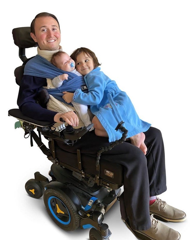 Quadriplegic father Josh Basile is pictured with his infant son and young daughter. His son is strapped to his chest with a sling and his daughter is wearing a blue bath robe and cuddling her younger brother as she sits on Josh's lap. Josh has light skin, short dark hair, and a big smile as he sits in his black power chair.