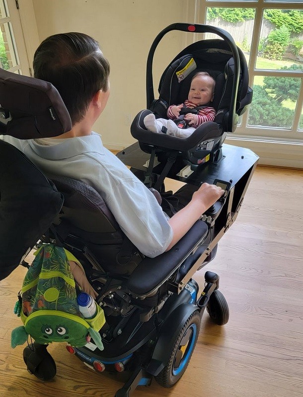 Seen from over Josh Basile's shoulder, infant son Calder sits in a black baby carrier that is hooked onto Josh's black power chair. From the back, Josh has short dark hair and light skin.