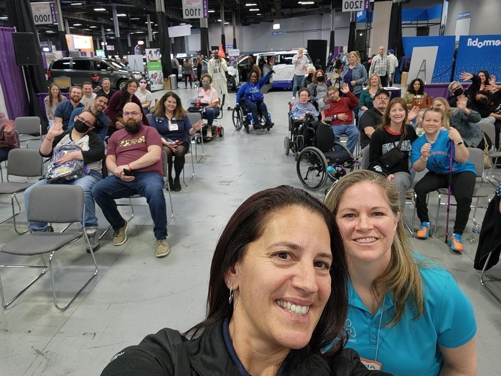 Kelly Green takes a selfie with Sonny Mullen at the Help Hope Live workshop at Abilities Expo NY Metro. Behind Kelly and Sonny are more than a dozen workshop attendees of different ages, ethnicities, and mobility levels. Some are in power chairs, using manual wheel chairs, or holding canes. Some wear masks.