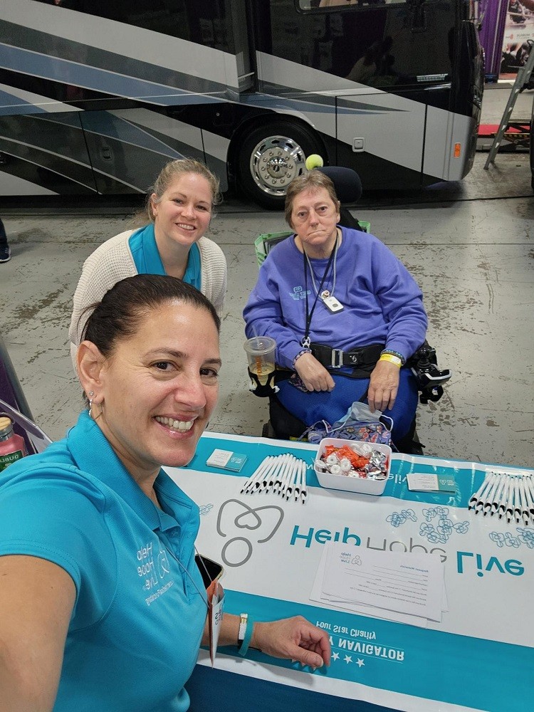 Kelly Green takes a selfie at Abilities Expo NY Metro with Sonny Mullen and client Jane Koza. Kelly has light skin, dark eyes, dark hair, and a teal Help Hope Live polo. Sonny has light skin, sandy hair, and a matching Help Hope Live polo. Jane has light skin, short hair, and dark eyes and is seated in her black power chair in front of the Help Hope Live table, which includes candy, pens, paperclips, and a banner.