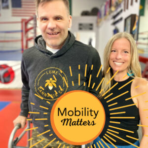 Spinal cord injury survivor Frank Guirlinger is pictured using a walker upright. He has light skin and gray hair. The woman next to him wears athletic gear and has light skin and blonde hair.