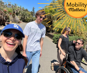 Megan, Arthur, and two friends are walking and rolling down a sidewalk with green vegetation and a clear blue sky visible behind them. Megan has a blue ballcap, sunglasses, light skin, and dark hair. Arthur is seated in his wheechair and has light skin, short gray-brown hair, and sunglasses. Their male and female companions both wear sunglasses and short-sleeved shirts. A graphic overlay reads Mobility Matters with a yellow and orange sunburst design.