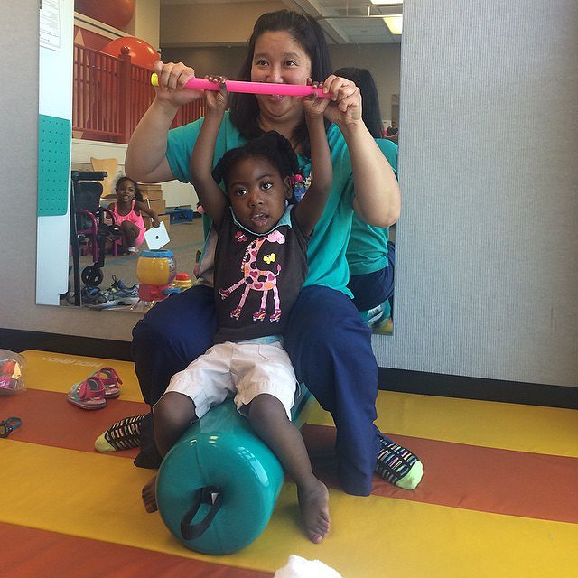 Zoë is in physical therapy seated on a teal cylindrical therapy cushion as she practices lifting a pink plastic bar over her head with assistance. Zoë has brown skin, dark brown eyes, textured black hair, and white shorts. Her physical therapy is Asian with straight black hair and brown eyes.