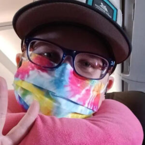 Shayn Pulley gives a peace sign as they sit in their airplane seat with a pink neck pillow. They have light skin, glasses, a ball cap, and a tie dye face mask.