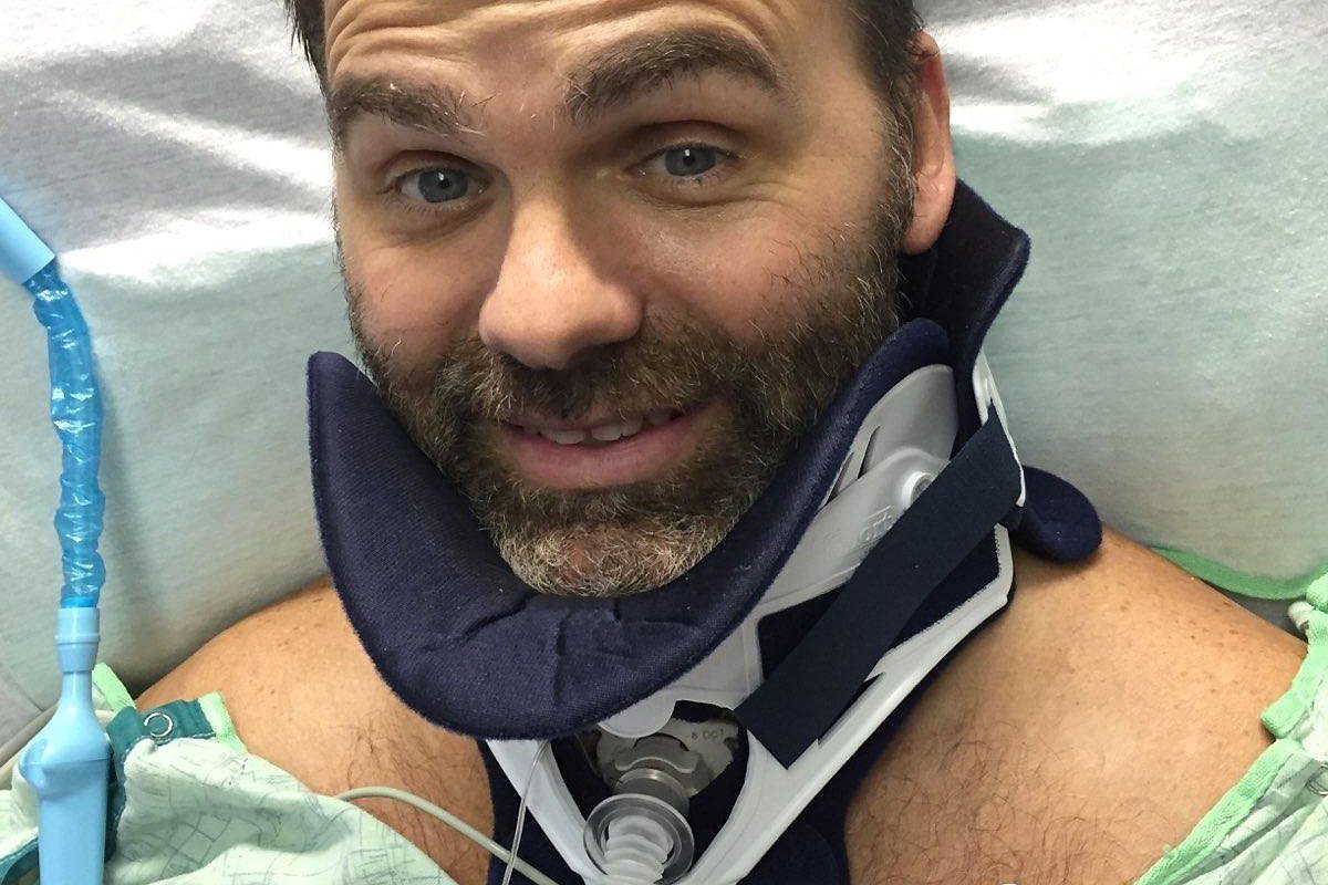 Frank Guirlinger is in the hospital after his spinal cord injury. He has light skin, gray hair, a short gray beard, and a neck brace and is wearing a hospital gown.