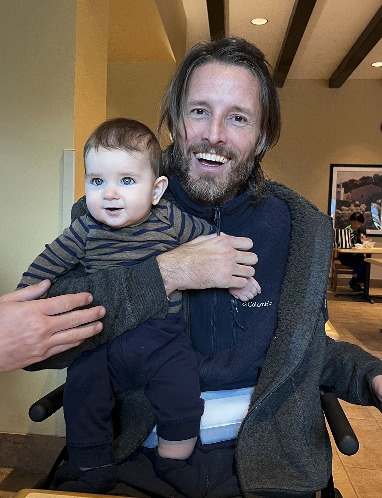Arthur grins joyfully as he holds an infant with light skin, striking blue eyes, and short brown hair. Arthur has light skin, brown hair to his shoulders, and a gray and brown beard and is seated in his wheelchair.