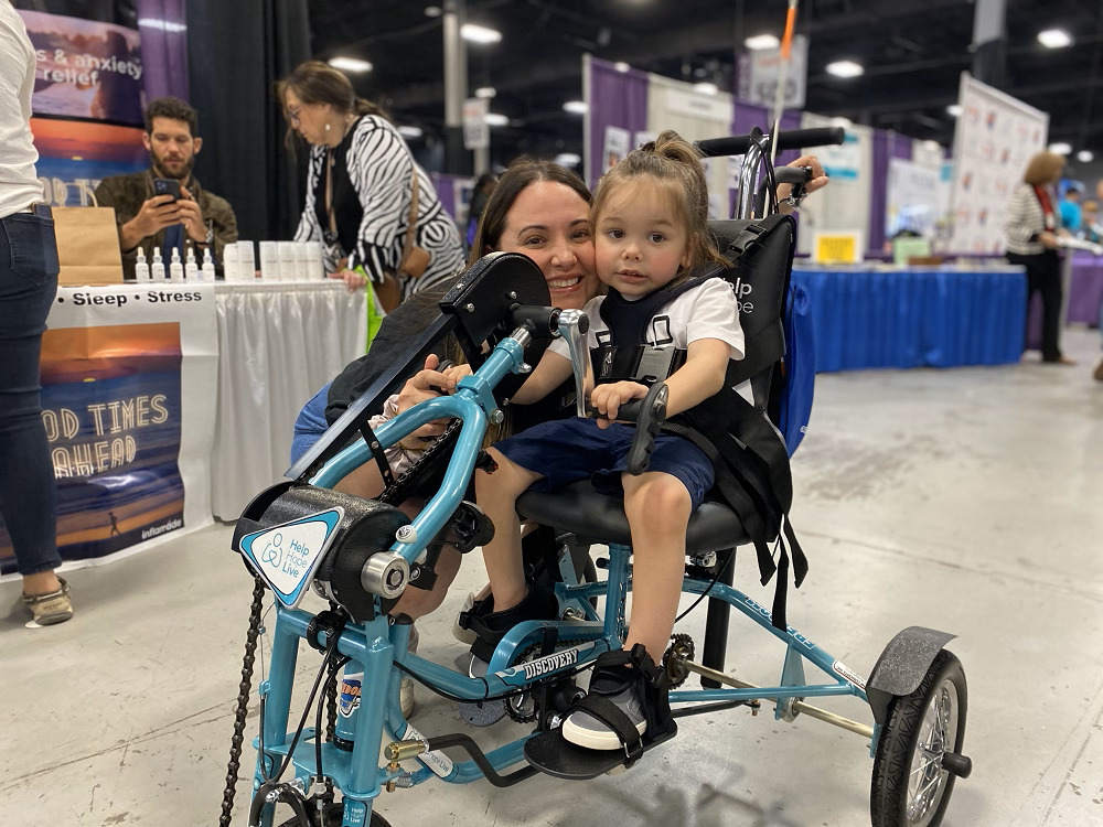 3-year-old client Easton Clark is strapped into his new Freedom Concepts Help Hope Live-themed adaptive tricycle. He has light skin, long light brown hair, and dark eyes. Mom is pictured kneeling with her face next to Easton’s. She has brown hair, brown eyes, and light skin. They are at Abilities Expo NY Metro.