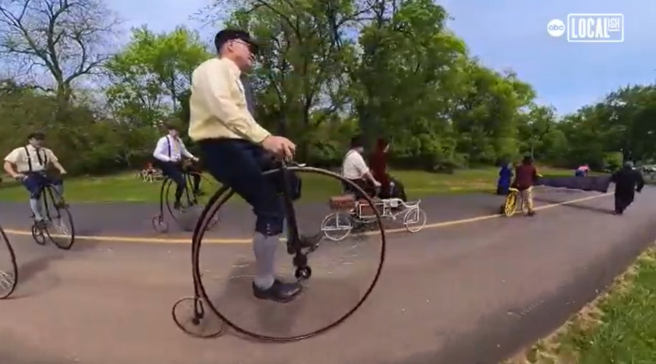 Antique bike riders on parade on a paved tree-lined road on a beautiful spring day. They wear old-fashioned garb like capri-length pants and caps and ride various antique bikes, including some seated bikes and some big-wheel bikes.