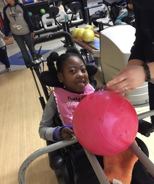 Zoë is ready to bowl seated in her black mobility device with a pink bib. She is receiving help to push a pink marbled bowling ball down a ramp in a bowling alley. Zoë has brown skin and brown eyes and textured black hair.