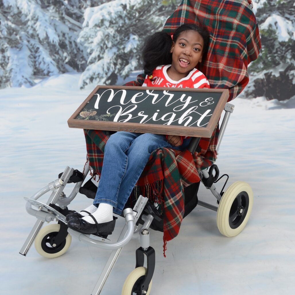 Zoë Sparks is ready for the season in a holiday portrait. Zoë has brown skin, brown eyes, and textured black hair. She wears jeans, black Mary Janes, and a red-and-white striped shirt and holds a sign that reads Merry & Bright. She is seated in her four-wheeled mobility device with a tartan blanket underneath her. Behind her is a snowy photography set.