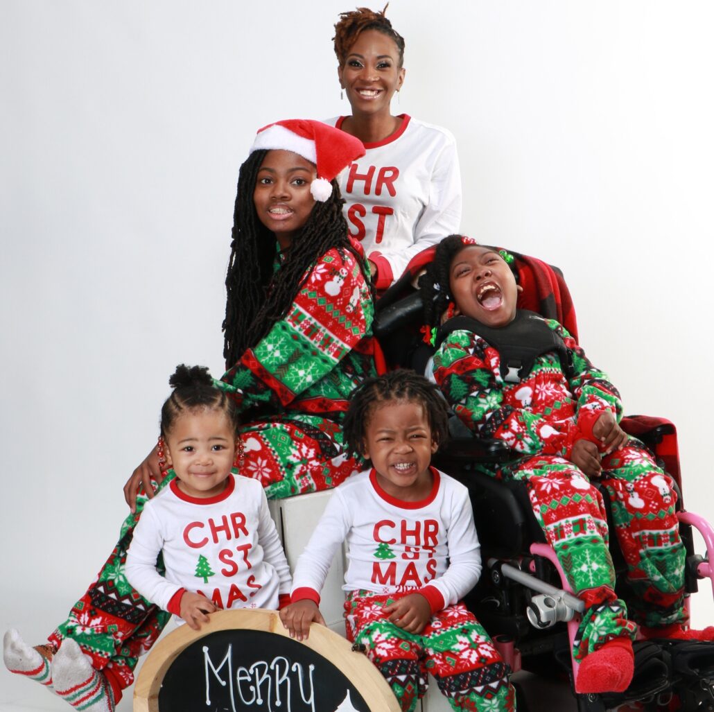 In a Christmas family portrait, mom Sierra Dillard and her four children smile in matching holiday-printed fleece pajamas. Sierra has short partially dyed locks, brown eyes, and brown skin. Her eldest daughter has long dark locks almost to her waist, brown skin, and brown eyes and wears a Santa hat. Zoë is strapped into her black mobility device and has brown skin, brown eyes, a huge smile, and textured black hair in braids. Their two youngest siblings have braided textured dark hair, brown skin, and brown eyes.