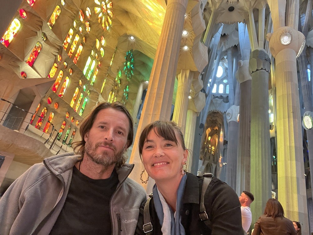 Arthur and Megan take a selfie in a beautiful cathedral church. Light is streaming in from vaulted ceilings past tall geometric columns and through intricate multicolored stained glass windows with rainbow gradients from red to blue. Megan has light skin, brown hair with bangs, and hoop earrings. Arthur has light skin, a short gray and brown beard, brown eyes, and brown hair to his shoulders.