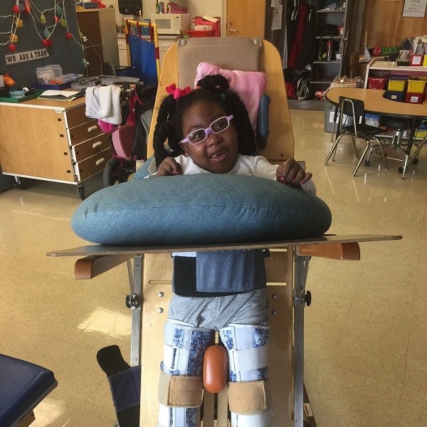 In a classroom setting, Zoë stands in a wood and metal standing frame with a fold-out desk and cushion in front of her. Zoë has brown skin, brown eyes, textured black hair in braids with a pink ribbon, and light pink glasses.