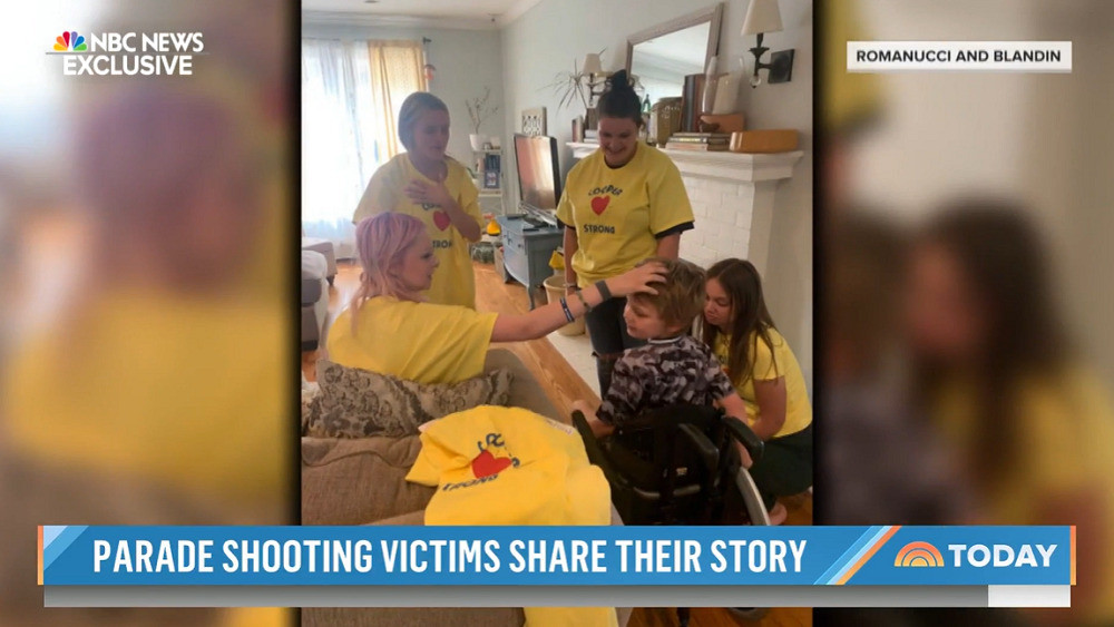 A screenshot of an NBC News Exclusive story. The headline ticker reads Parade Shooting Victims Share Their Story. The photo credit is Romanucci and Blandin. The photo is four women in yellow COOPER STRONG t-shirts. One of the women places a hand on Cooper's forehead. Cooper is a child with light skin and sandy short hair seated in a black wheelchair.