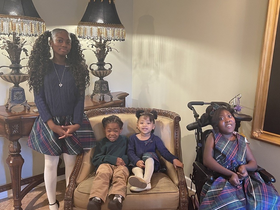 Zoë and her siblings are in matching outfits for a family portrait. Zoë has brown skin and brown eyes and wears a tartan dress as she sits in her black mobility chair. Her textured hair is partially curled. Her older sister stands with a blue ribbed sweater and a tartan skirt with a silver locket around her neck and solid white tights. Her sister has brown skin and brown eyes and curly brown hair in two long pigtails. Zoë's youngest siblings are a boy and a girl wearing formal attire with tartan accents.