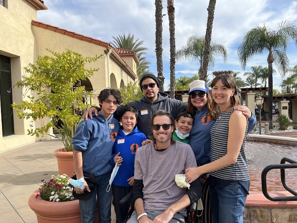 Arthur, Megan, and a group of friends are outside in a beautiful courtyard with a fountain, palm trees, and cloudy blue skies. Their friends appear to be part of the same family with light brown skin, dark hair, and matching hoodies. The woman and man wear sunglasses and three children vary in age beside them. Megan has light skin, shoulder-length brown hair with bangs, and a short-sleeved navy and white striped top and jeans. Arthur wears a t-shirt over a long-sleeved shirt, sunglasses, and a hospital bracelet and has light skin, brown shoulder-length hair, and a short gray and brown beard.