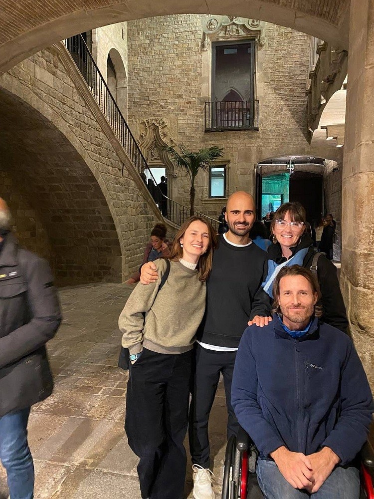 Arthur, Megan, and two friends stand in a striking stone courtyard with decorative doorways and archways. Arthur is seated in his wheelchair and has light skin, brown hair to his shoulders, brown eyes, and a short gray-and-brown beard. Megan has dark hair with bangs, glasses, light skin, and hoop earrings.