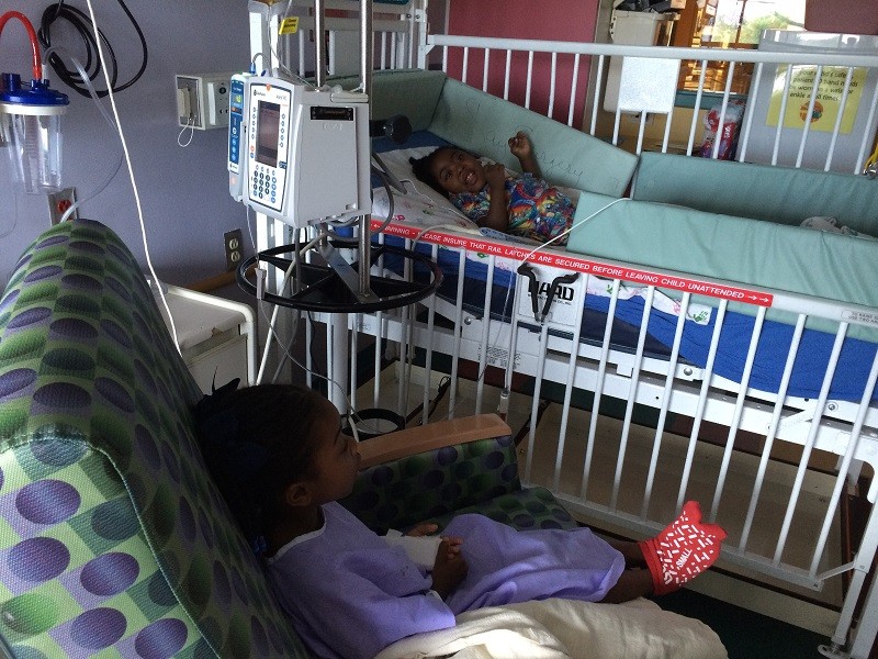 Zoë lies in a hospital bed in a multi-colored gown with a smile on her face. Near her sits a sibling in a lavendar hospital gown and hospital socks. Both girls have brown skin and textured black hair.