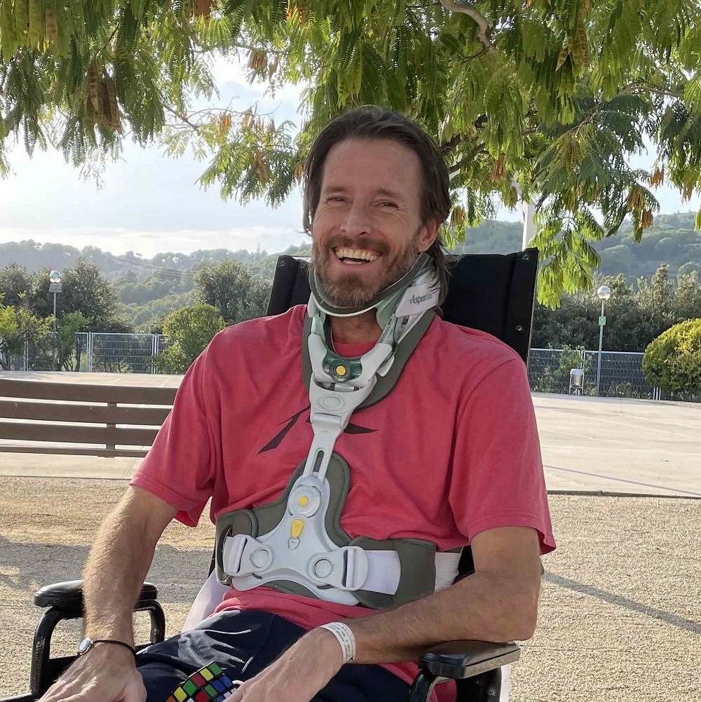 Arthur is grinning and looks like he has just been laughing as he sits outside in his wheelchair with a brace visible around his neck, down his torso, and across his lower chest. He has light skin, a short gray-brown beard, and dark hair to his shoulders.