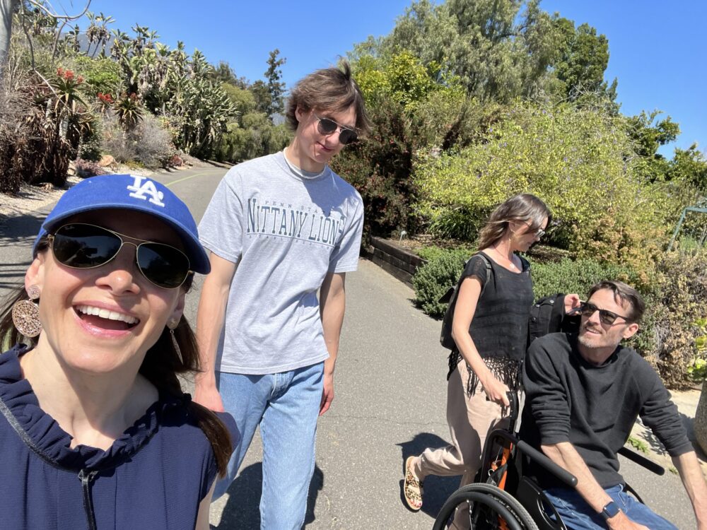 Megan, Arthur, and two friends are walking and rolling down a sidewalk with green vegetation and a clear blue sky visible behind them. Megan has a blue ballcap, sunglasses, light skin, and dark hair. Arthur is seated in his wheechair and has light skin, short gray-brown hair, and sunglasses. Their male and female companions both wear sunglasses and short-sleeved shirts.