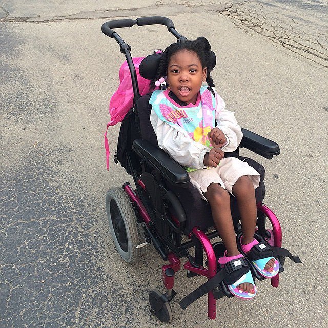 Zoë is outdoors on a blacktop seated in a black push-able wheelchair. Zoë has brown skin, textured black hair in braids, and brown eyes. In soft sandals, her toenails are painted pink.