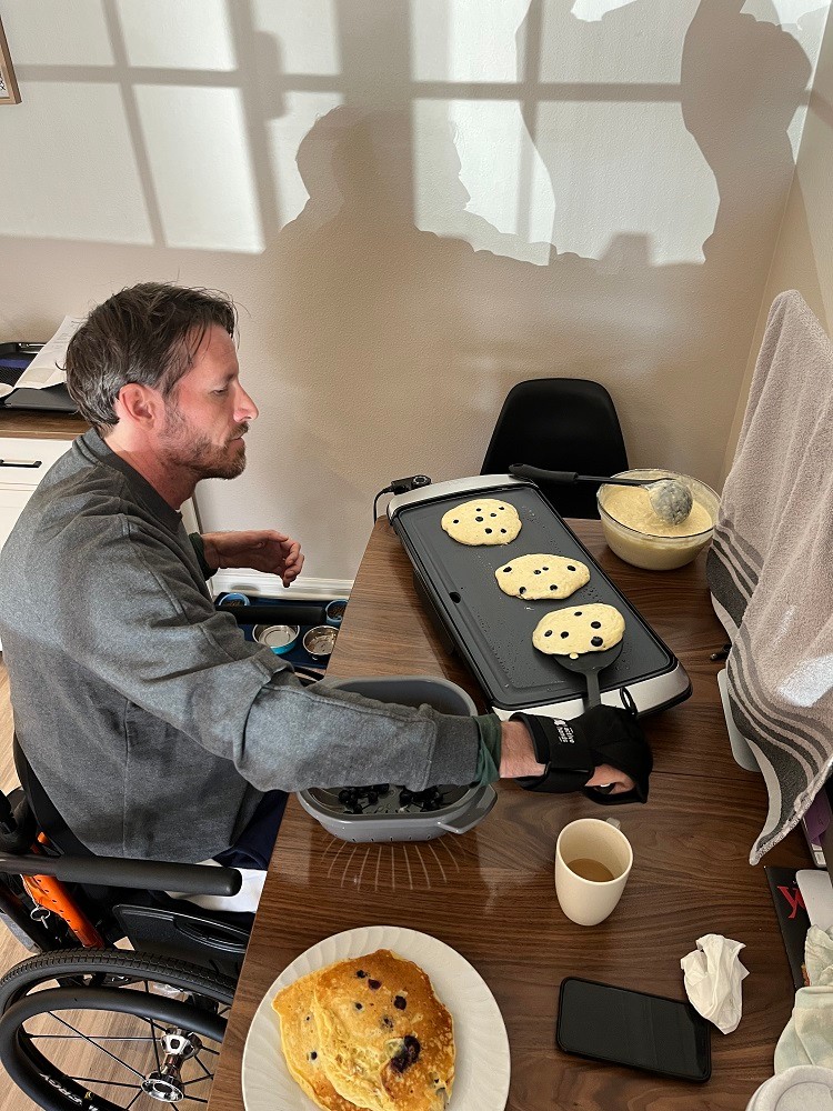 Arthur flips three chocolate chip pancakes that are cooking on a griddle while seated in his wheelchair. Arthur has light skin, short dark hair, and a short beard.