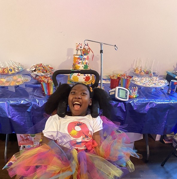 Zoë is overjoyed at her birthday party. She sits in her black mobility and has brown skin, brown eyes, and textured hair in two pigtails. She wears a t-shirt with the number 9 and a multicolored tulle tutu. Behind her is a table full of rainbow-colored treats and a Happy Birthday cake covered in painting decorations and edible paint splatters.