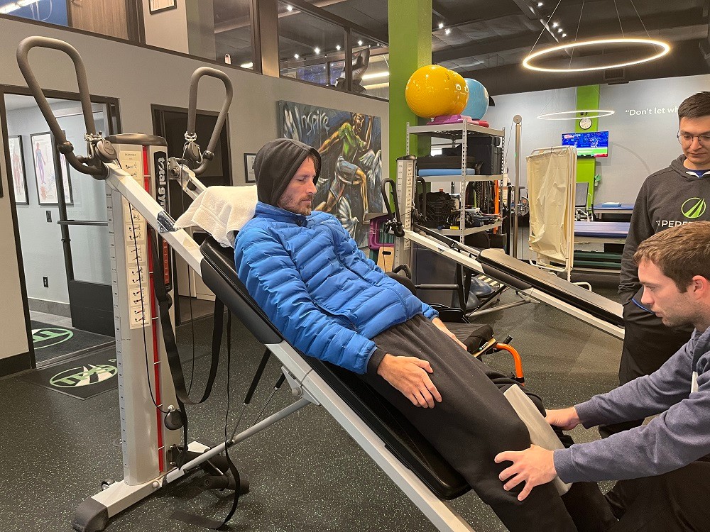 Arthur is in a rehab or physical therapy setting with equipment and trainers visible behind him. He is lying on an inclined piece of equipment with his arms by his sides and lifting his head with his eyes closed in concentration as a trainer holds his legs. He wears a gray hoodie with the hood pulled over his head and a blue quilted warm jacket, and he has light skin a short beard.