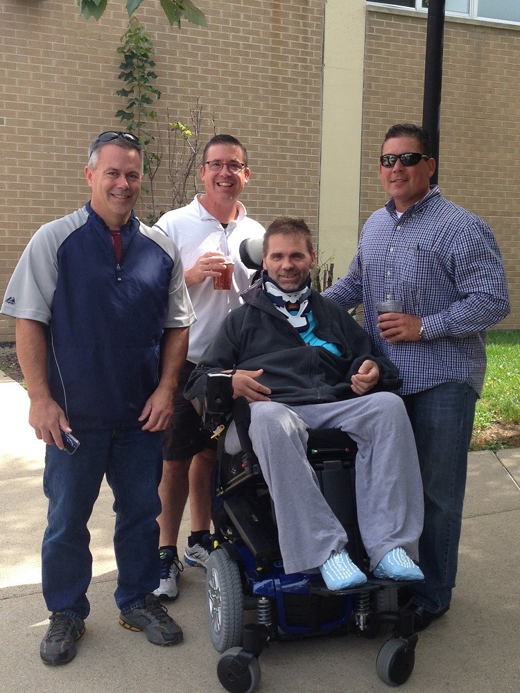 Frank is seated in his black power chair with a brace around his neck. He has light skin and gray hair. Three men stand beside his chair and smile. They are outside of a rehabilitation facility.