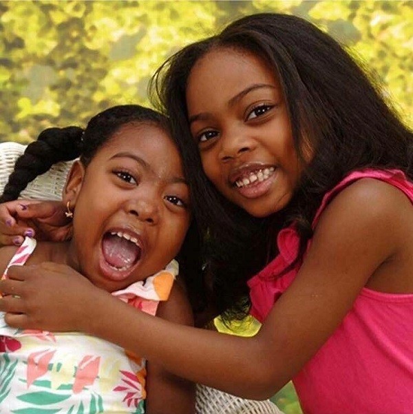 Zoë receives a hug from her older sister as both girls look at the camera and smile. Zoë and her sister both have brown skin, brown eyes, and textured dark hair (Zoë's hair is in two bubble braids).