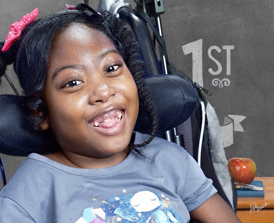 Zoë gives a beautiful smile with her tongue resting between her lips in a 1st grade photo portrait from Lifetouch. Zoë has brown skin, dark brown eyes, and textured black hair that is partially braided and includes two pink polka-dotted bows.