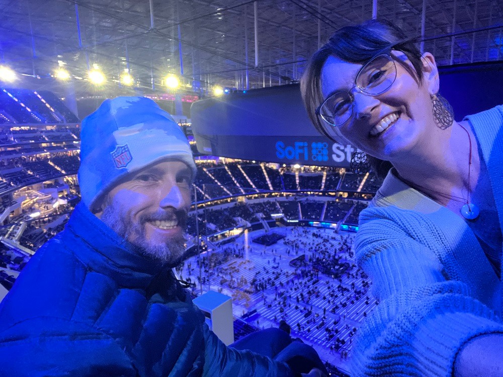 Arthur and Megan take selfie at an event at SoFi stadium from high in the stadium seating. Arthur has light skin, a short beard, and a beanie with a warm quilted jacket. Megan has light skin, glasses, dark hair pulled back, and a white ribbed sweater.