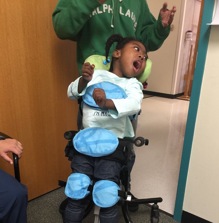 Zoë stands in a black mobility device. She is strapped in at the chest, waist, and knees, and the device can roll. Zoë has brown skin, textured black hair, and brown eyes.