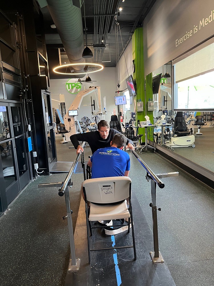 Arthur is in a physical therapy or rehabilitation facility with equipment and trainers visible in the background. A seated trainer helps him to grip parallel bars and lift himself into a standing position. Arthur has light skin, brown hair, and a short beard.