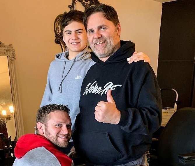 Spinal cord injury survivor Frank Guirlinger stands with assistance and gives a thumbs-up sign. He has light skin, a light gray short-cropped beard, and dark hair. He stands beside a young adult as a man helps to stabilize his legs. All are smiling.