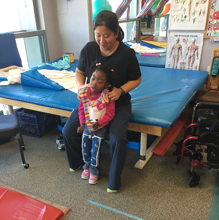 Zoë participates in physica therapy in a rehab setting. Her PT supporter is seated on a blue therapy table to support Zoë's body as she stands. Zoë has brown skin, textured black hair, a Hello Kitty sleeveless top over a pink long-sleeved shirt, jeans, soft leg braces, and pink sneakers.