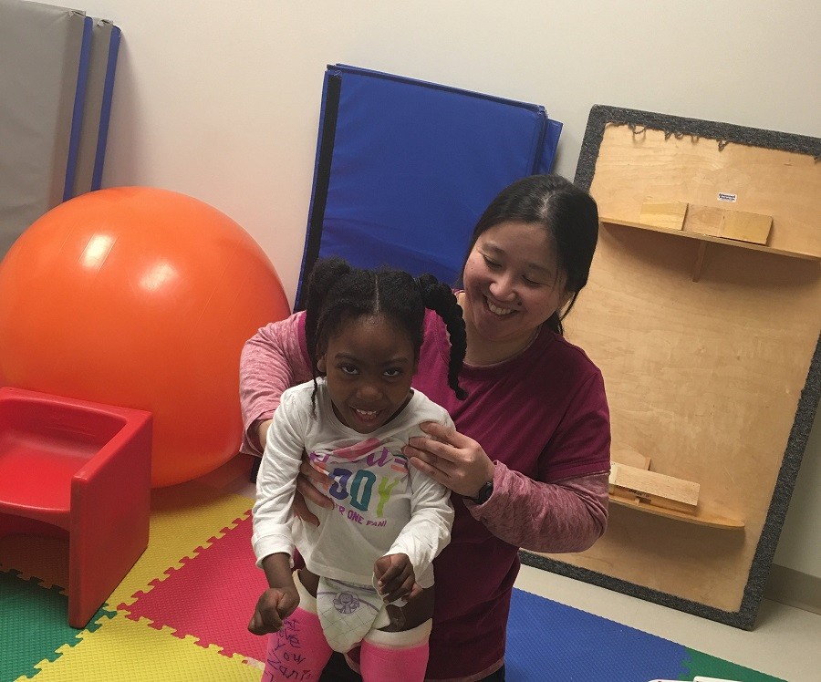 Zoë looks excited as she participates in physical therapy. She has brown skin, brown eyes, and textured black hair. She is standing with assistance wearing a long-sleeved white t-shirt, diaper, and two bright pink leg casts. Her PT professional is Asian with straight black hair and a big smile, wearing a mulberry-colored t-shirt.