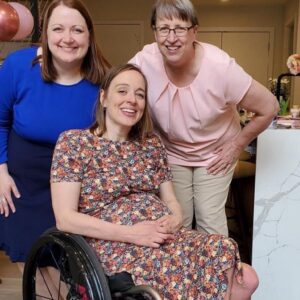 Cindy Kolbe leans down for photo with her daughter, Beth, at a baby shower. Cindy has light skin, glasses, short gray hair, and a pink blouse. Next to her, a smiling woman has shoulder-length red hair, light skin, and a blue dress. Cindy daughter Beth is seated in her black manual wheelcahir and wears a flower print dress. She has light skin, shoulder-length brown hair, light eyes, and a growing baby bump.