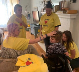 Four women in a living room wearing yellow COOPER STRONG t-shirts. One of the women places a hand on Cooper's forehead. Cooper is a child with light skin and sandy short hair seated in a black wheelchair.